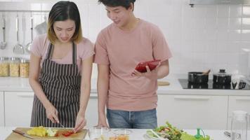Happy Asian beautiful young family couple husband and wife cooking vegetable salad in kitchen together at home. The man and woman according online cooking class to recipe on laptop computer video