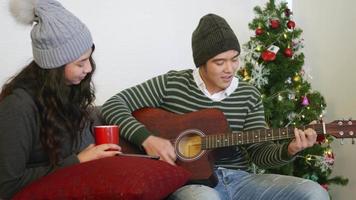 Happy Asian beautiful young family couple husband and wife on sofa great time in home living room, man and woman sit sofa playing guitar singing song winter holidays together enjoying, slow motion video