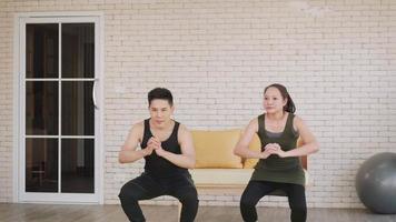 Happy Asian beautiful lifestyle family couple doing YOGA standing on exercise sitting squat position workout at home together on mat. Two people sport healthy concept. Slow motion video