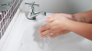 Primer plano de las manos de una mujer joven usando jabón y lavándose las manos bajo el grifo de agua, las mujeres se lavan las palmas con burbujas blancas en el lavabo del baño del hotel para protegerla de la covid-19 video