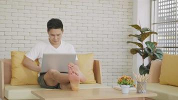 feliz homem bonito asiático usando e trabalhando com um laptop com internet online de casa. jovem sorrindo lê a tela do computador enquanto relaxa no sofá. câmera lenta video