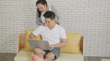 Happy Asian family couple husband and wife laughing sitting on sofa using laptop computer working from home Business man sitting on sofa home working on laptop her woman is giving her shoulder massage