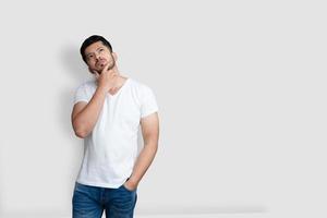 Hombre guapo asiático en camiseta blanca que tiene dudas mientras mira hacia arriba sobre fondo blanco aislado foto