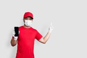 Repartidor empleado en gorra roja camiseta en blanco uniforme máscara facial mantenga negro aplicación de teléfono móvil aislado sobre fondo blanco.