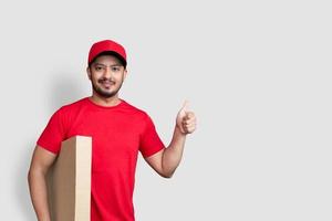 Repartidor empleado en gorra roja camiseta en blanco pulgar arriba uniforme mantenga caja de cartón vacía aislado sobre fondo blanco. foto