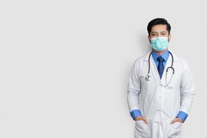 Young male doctor face covered with mask and hands in coat pocket over isolated background, health concept photo