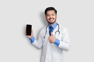 Doctor Holding Phone Showing Thumb Up Isolated. Pakistani Man Doctor Technology Medicine at Home. Phone clear Screen. photo