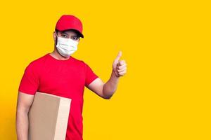 Delivery man employee in red cap blank t-shirt uniform face mask hold empty cardboard box isolated on yellow background