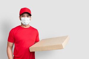 Delivery man employee in red cap blank t-shirt uniform face mask hold empty cardboard box isolated on white background photo