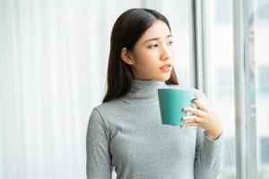 Mujer asiática bebiendo café por la ventana durante las vacaciones