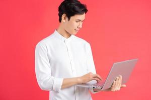 Una foto de un apuesto hombre asiático de pie sobre un fondo rojo con una computadora