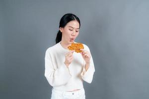 Joven asiática vistiendo un suéter con una cara feliz y disfruta comiendo pollo frito sobre fondo gris
