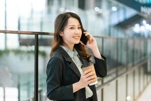 Retrato de una joven directora asiática de pie, tomando café y escuchando el teléfono foto