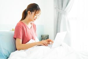 Joven mujer asiática sentada en su cama por la mañana y usando la computadora portátil para el trabajo