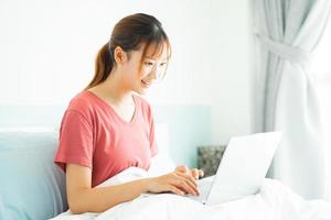 Joven mujer asiática sentada en su cama por la mañana y usando la computadora portátil para el trabajo foto