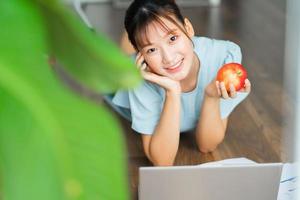 Joven mujer asiática sosteniendo una manzana y usando su computadora portátil en la mañana