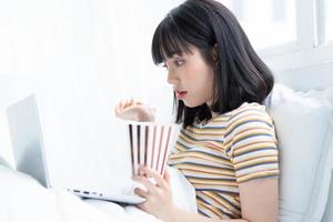 Young asian woman using laptop to watching tv shows, eating popcorn at home photo