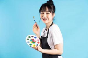 Beautiful Asian female painter holding quill pen and color palette on blue background photo