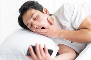 The young man is lying on the bed yawning, still holding the phone photo