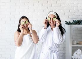 Dos hermosas mujeres haciendo procedimientos de spa cubriéndose los ojos con pepinos foto