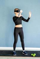 Young blonde woman in sport clothes wearing virtual reality glasses standing at fitness mat using VR interactive menu photo