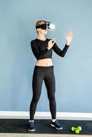 Young blonde woman in sport clothes wearing virtual reality glasses standing at fitness mat using VR interactive menu