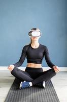 Young blonde woman in sport clothes wearing virtual reality glasses meditating on fitness mat