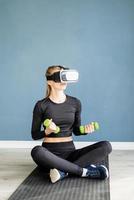 Young blonde woman in sport clothes wearing virtual reality glasses sitting on fitness mat