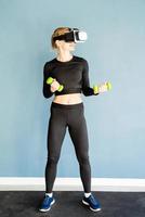 Young blonde woman in sport clothes wearing virtual reality glasses standing at fitness mat working out with dubbells photo