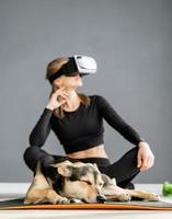 Young woman in sport clothes wearing virtual reality glasses sitting on fitness mat with dog photo