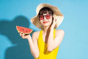 Beautiful Asian woman wearing yellow jumpsuit on blue background and eating tropical fruits, summer concept photo