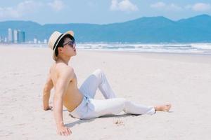 Barebacked young Asian man sitting on the sand and looking at the sea