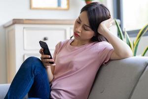 Beautiful young Asian woman with a sad expression sitting using the phone on the sofa at home photo