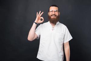 Foto de hombre guapo con barba en camiseta blanca mostrando gesto ok sobre fondo negro