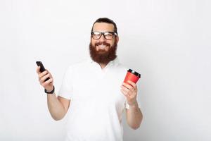 Cheerful bearded man in white t-shirt using smartphone and holding cup of coffee to go
