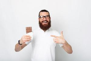 Cheerful young bearded man smiling and pointing at bar of chocolate photo