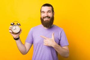 Portrait of handsome young man with beard pointing at alarm clock and smiling