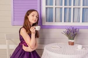 A teenage girl in a purple dress at home or in a cafe. The girl is drinking tea from a white cup. photo