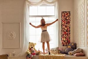 retrato de una mujer joven con un vestido blanco enderezar cortinas blancas claras junto a la ventana. foto