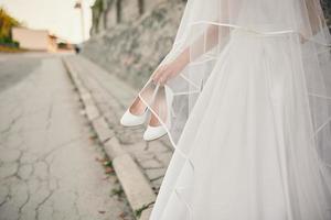 la novia con un vestido de novia y un velo camina por la calle con los zapatos en la mano foto
