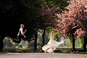 novios felices se divierten en el parque floreciente de la primavera de Sakura. el hombre con barba está saltando, la mujer con vestido largo está bailando. pareja de recién casados en el parque. recién casados. boda rústica. foto
