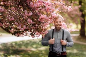 Bearded man with stylish haircut with sakura flowers on background. Hipster in in suspenders near with branch of sakura flowers in teeth. Harmony with nature concept. photo