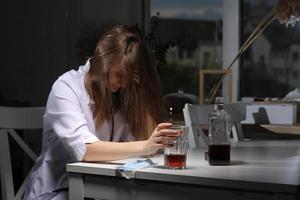 Doctora sentada en la mesa y sosteniendo un vaso con whisky o coñac en casa después de un arduo trabajo, mujer deprimida que bebe alcohol fuerte y sufre problemas de coronavirus. foto