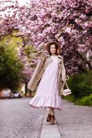 young beautiful stylish woman in hat and pink dress walking near sakura flowers in the park. Spring concept. Uzhhorod, Ukraine photo