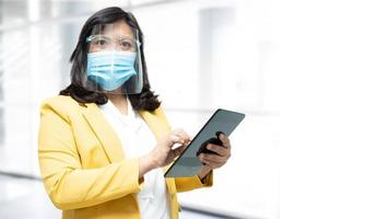 Asian lady holding tablet and wearing mask new normal in office for protect safety infection Covid-19 Coronavirus with copy space. photo