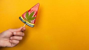 Hermosa piruleta roja dulce con marihuana en mano sobre fondo amarillo, dulces con cannabis foto