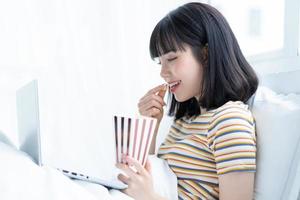 Young Asian woman using laptop to watching tv shows, eating popcorn at home photo