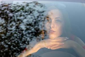 una mujer joven y hermosa con el pelo largo se sienta al volante del automóvil y mira soñadora a través del resplandor del parabrisas delantero. foto