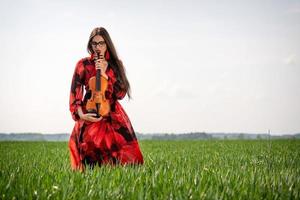 Young woman in red dress with violin in green meadow - image photo