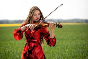 Young woman in red dress playing violin in green meadow - image photo